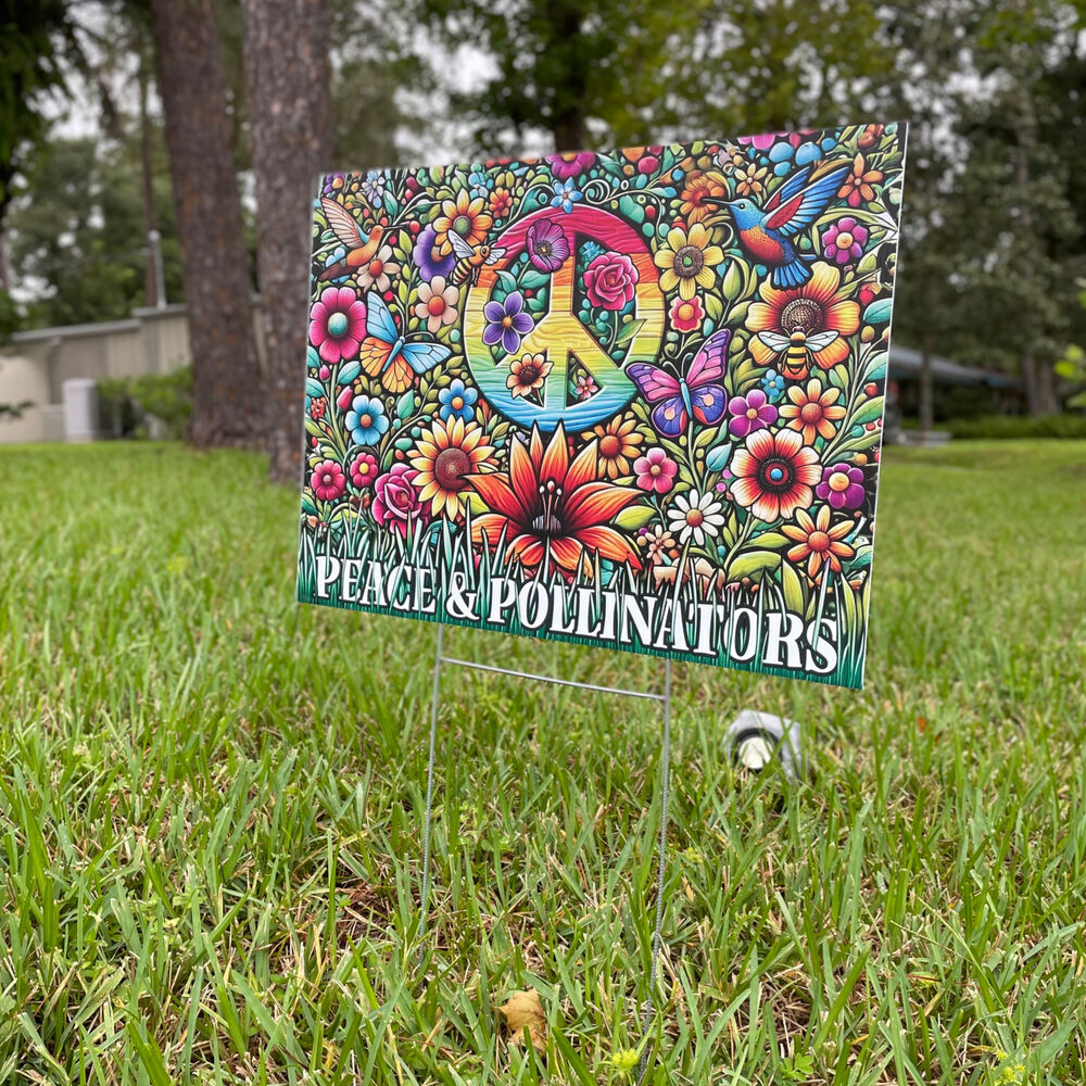 The printed Peace & Pollinators sign on a wire stake in a green lawn, trees behind it.
